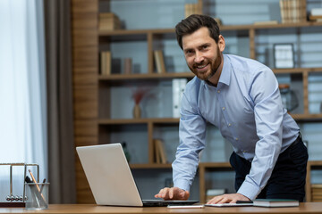 Obraz premium Smiling mature businessman in business suit engages in work on his laptop amidst a disorganized home office environment.