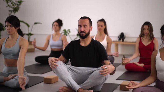Group of women and man with eyes closed breathing sitting cross-legged in lotus flower posture on mats during yoga class, practicing meditation improving self-awareness. Wellness lifestyle concept.