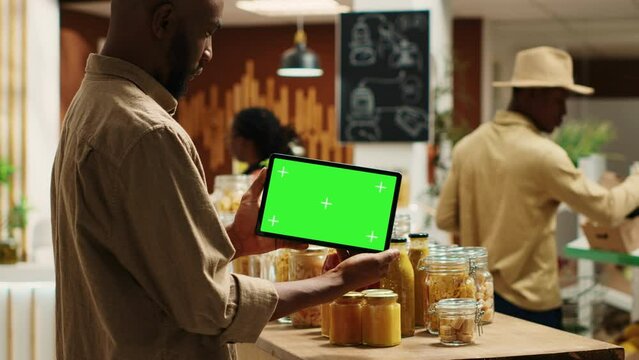 African american buyer shows tablet with greenscreen template in local grocery store, looking at blank copyspace next to chemicals free items. Regular client holds device with chromakey. Camera A.