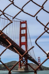 A vertical shot of the Golden Gate Bridge visible through a rusty fence at Fort Point