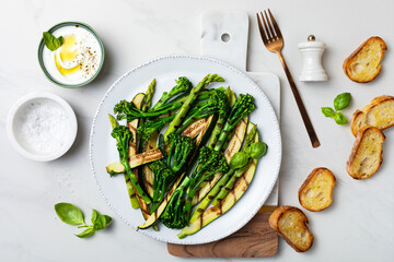 Grilled green spring vegetables: zucchini, asparagus, broccolini with yoghurt sauce on white marble background.