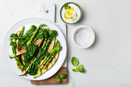 Grilled green spring vegetables: zucchini, asparagus, broccolini with yoghurt sauce on white marble background. Top view, copy space. .