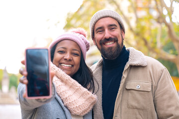 Happy multiracial young couple smiling pointing to empty smartphone screen outdoors