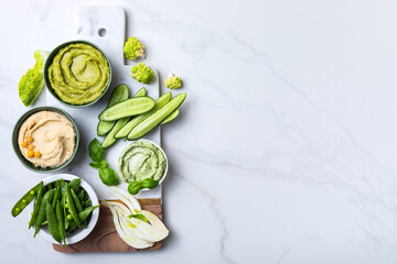 Green spring vegetable appetizer platter with dips. Healthy crudites snack board on white marble background.