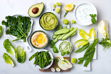 Green spring vegetable appetizer platter with dips. Healthy crudites snack board on white marble...