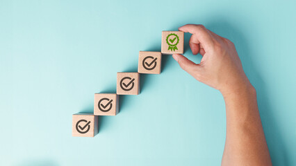 A hand is placing the final wooden block with a green certificate checkmark, symbolizing achievement, quality assurance and completion, against a plain pastel blue background. - obrazy, fototapety, plakaty