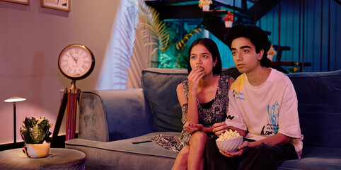 Indian Asian teen girl boy couple pair sitting sofa couch watch horror film movie TV enjoy fun joy late night indoor home gen z male female man lady friend eat snack hold bowl look scared action video