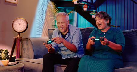Indian Asian Hindu happy smiling old aged cute gen x couple elder older male female man lady sitting cozy sofa couch play video game hold gadget use device enjoy fun joy late night room home house © Raushan_films