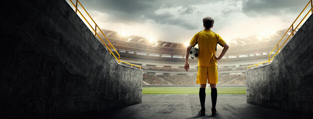 Young guy, soccer player in yellow uniform standing with football ball and looking at 3D stadium....