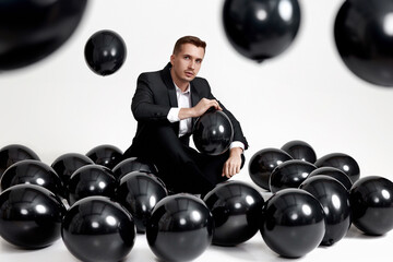 elegant caucasian man in suit tuxedo sitting on studio floor with a lot of black air balloons