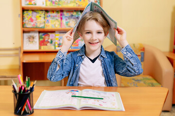 boy 7 years old in a school building, sitting at a desk, holding textbooks in his hands, covering...