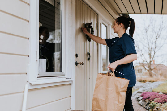In-home nurse carrying paper bag with shopping and knocking to entrance door