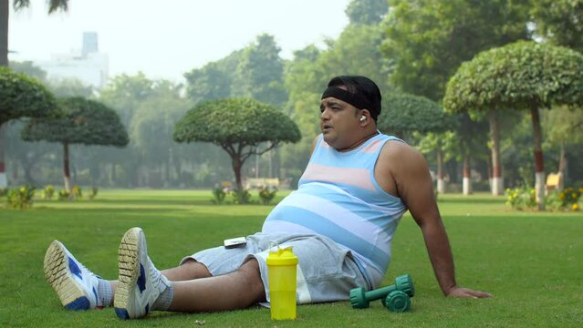 An Indian man listening and vibing to a song in the playground - relax-position  healthy-lifestyle  tired after workout. A man sitting and relaxing after a tiring workout - green park  relaxing  ov...
