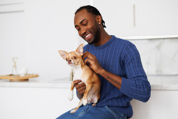 Smiling young african american man in jumper holding chihuahua dog in kitchen