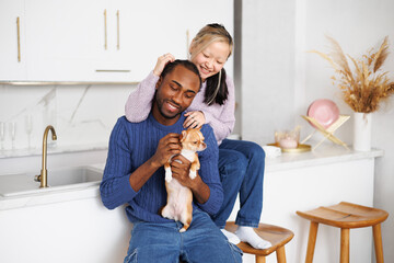 Smiling asian woman in sweater hugging african american boyfriend with chihuahua dog in kitchen
