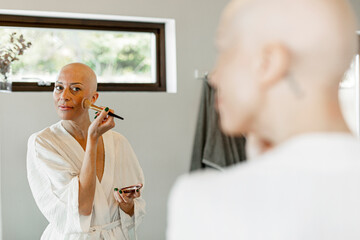 Reflection of bald woman with cancer applying blusher on cheek at home