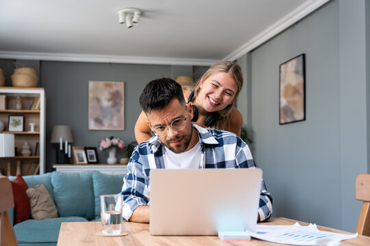 Young nervous college student man trying to learn and prepare for final exam while his girlfriend teasing him and making fun, trying to relax him. Freelance couple, job search male working on laptop