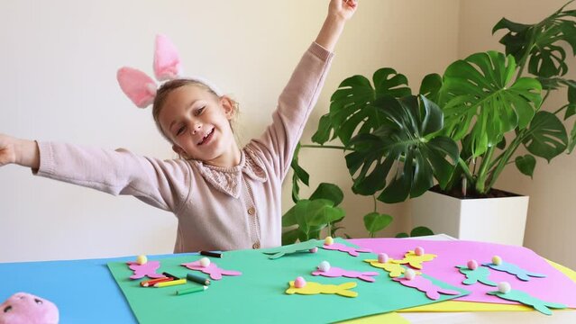 Child girl in rabbit ears make Easter paper colored garland Easter bunny. Cute children's crafts for Easter party at home. Holiday Art Activity Kid. Fine motor skills, creativity and hobby DIY