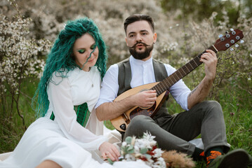a bearded groom plays a stringed instrument and a girl sits in a spring meadow