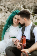 a bearded groom plays a stringed instrument and a girl with sits in a spring meadow