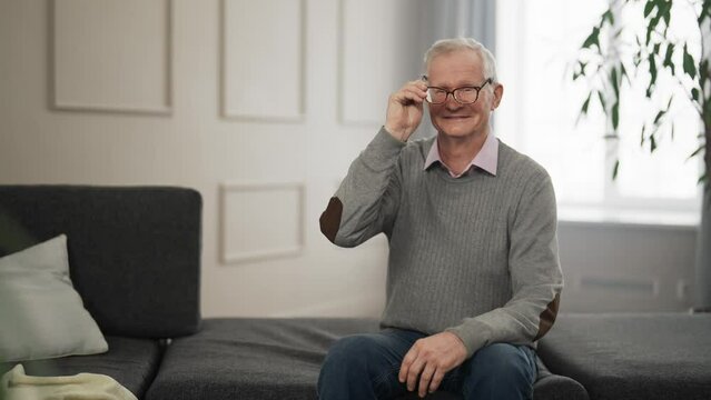 Senior retired man in glasses at home. Old gray haired intelligent male sits on couch at home smiling, watching TV or video. Entertainment, leisure pastime resting relaxing on weekend concept.