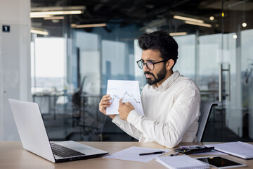 Serious and focused young indian man sitting at desk and showing documents with graphs and...