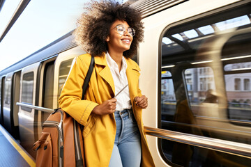 Cheerful smiling African American woman traveler hurries to board a high-speed train on a modern railway platform, ready to go a new trip. Concept of eco-friendly, modern public transport. Copy space - obrazy, fototapety, plakaty