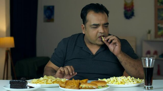 A middle-aged unhealthy hungry man enjoying a variety of snacks at home - munching time  junk food  junk snacks. An Indian obese man eating chips  samosas  popcorn  and drinks -  hungry man  bad ch...