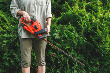 Woman holding electric hedge trimmer. Gardener is ready for trimming overgrown bushes at backyard. Gardening equipment