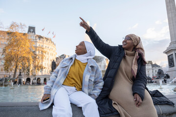 UK, London, Young female tourists in hijabs relaxing in city square - obrazy, fototapety, plakaty