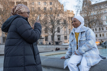 UK, London, Young woman in hijab photographing friend sitting on fountain