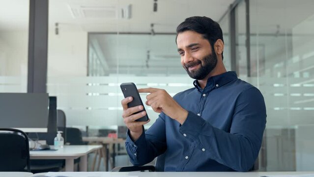Happy busy Indian business man executive using mobile phone working in office. Professional businessman employee looking at smartphone checking banking apps typing on cell sitting at workplace desk.