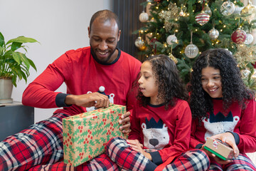 Smiling father with two daughters unpacking gifts in front of Christmas tree