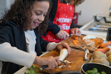 Girl peeling parsley root in kitchen, mother in background - obrazy, fototapety, plakaty