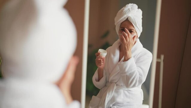 Relaxed senior woman applying clay face mask for moisturizing skin after bath. Satisfied old lady in white bathrobe. Skin care, home cure beauty spa cosmetology, domestic procedures treatment concept.