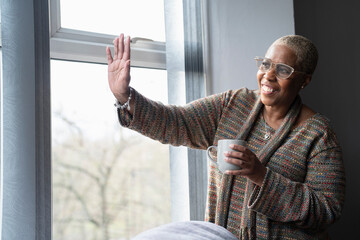 Mature woman with coffee cup looking through window and waving