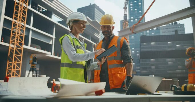 Construction and Site Managers Having a Meeting at a Construction Site with Skeleton Frame Building with Concrete and Steel Beams in the Background. Specialists Using Tablet to Review the Blueprints