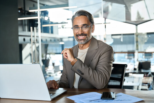 Happy middle aged 50 year old professional business man, smiling older executive ceo manager, smiling entrepreneur wearing suit sitting at desk in office working on laptop looking at camera, portrait.