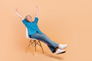 Full length photo of good mood retired person wear blue shirt jeans sitting on chair hands up relaxing isolated on beige color background