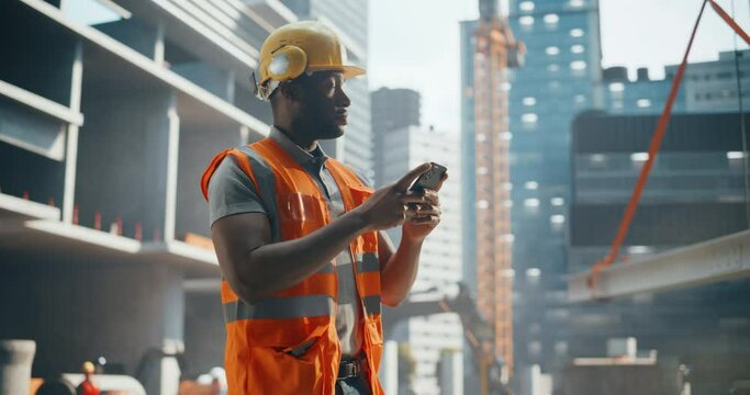 Portrait of Handsome African Builder, Worker, Craftsman Wearing a Hard Hat and a Vest while Standing at a Commercial Building Construction Site and Using a Smartphone to Browse Through Social Media