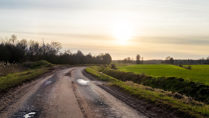 The image captures a serene rural scene, featuring a winding country road meandering through the landscape and leading into the soft glow of the setting sun. The road is flanked by green fields and