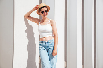 Beautiful smiling model in sunglasses. Female dressed in summer hipster white T-shirt and jeans. Posing near white wall in the street. Funny and positive woman having fun outdoors, in hat, sunglasses