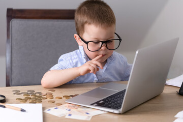A little boy in glasses sits at a table and looks at a laptop screen. On the table next to the laptop are money, coins and banknotes. A boy sits on a chair at the table, does his homework and counts