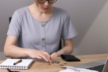 A woman with glasses counts money, coins, cents. On the table is a notebook with blank sheets, a pen, a magnifying glass and a laptop. Poverty, economy, finance. High quality photo