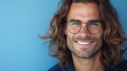 Obraz premium A man with long curly hair wearing glasses smiling against a blue background.