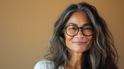 Obraz premium A woman with long wavy gray hair wearing glasses smiling gently against a warm blurred background.