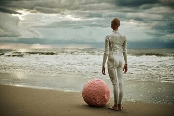 Hairless performer girl with alopecia in white futuristic suit stands on sea beach with pink sphere...