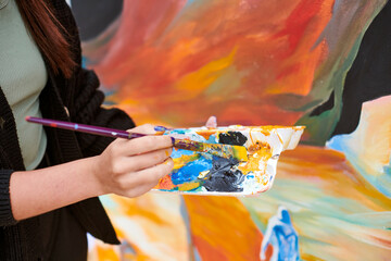 Female painter hand dips paintbrush into palette of colorful paints for live painting of picture...