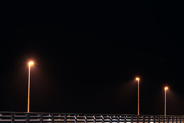 Street lampposts casts dim yellow warm along shore pier standing against pitch black canvas of...