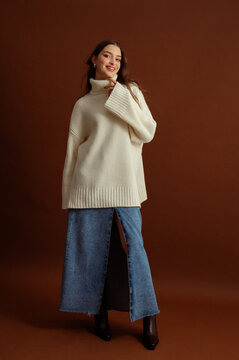 Fashionable happy smiling woman wearing oversized white turtleneck sweater, maxi blue denim skirt, pointed toe ankle leather boots, posing on brown background. Full-length studio fashion portrait